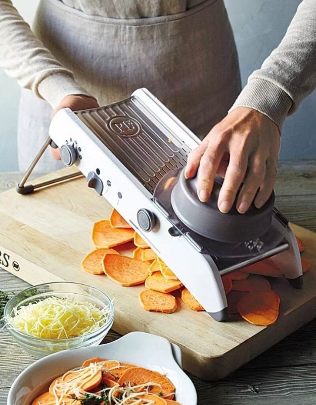 Person using a mandoline slicer to slice sweet potatoes on a wooden cutting board. Adjustable mandoline slicer slicing sweet potatoes with hand guard.